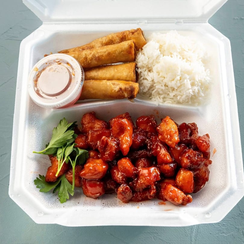 Sweet and sour chicken, white rice, egg rolls, and sauce in a white takeout container.