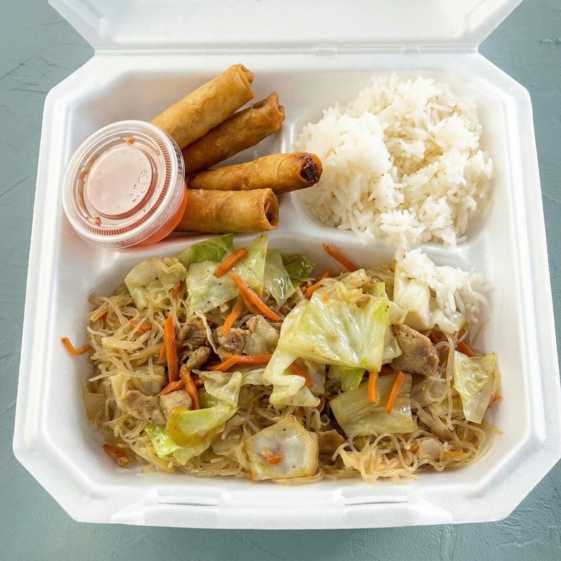 A takeout container with pancit, white rice, four lumpia rolls, and a small cup of sauce.
