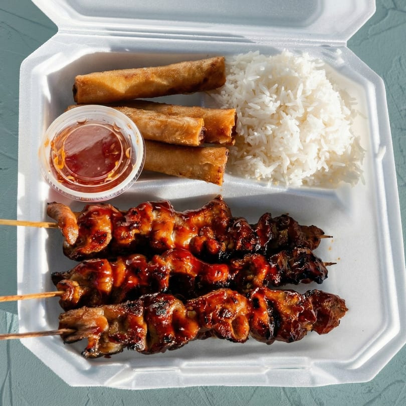 Takeout container with three chicken skewers, three egg rolls, white rice, and a side of sauce.