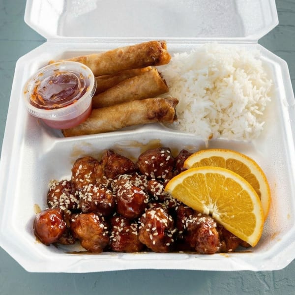 Orange chicken with sesame seeds, white rice, egg rolls, and sweet and sour sauce in a takeout container.