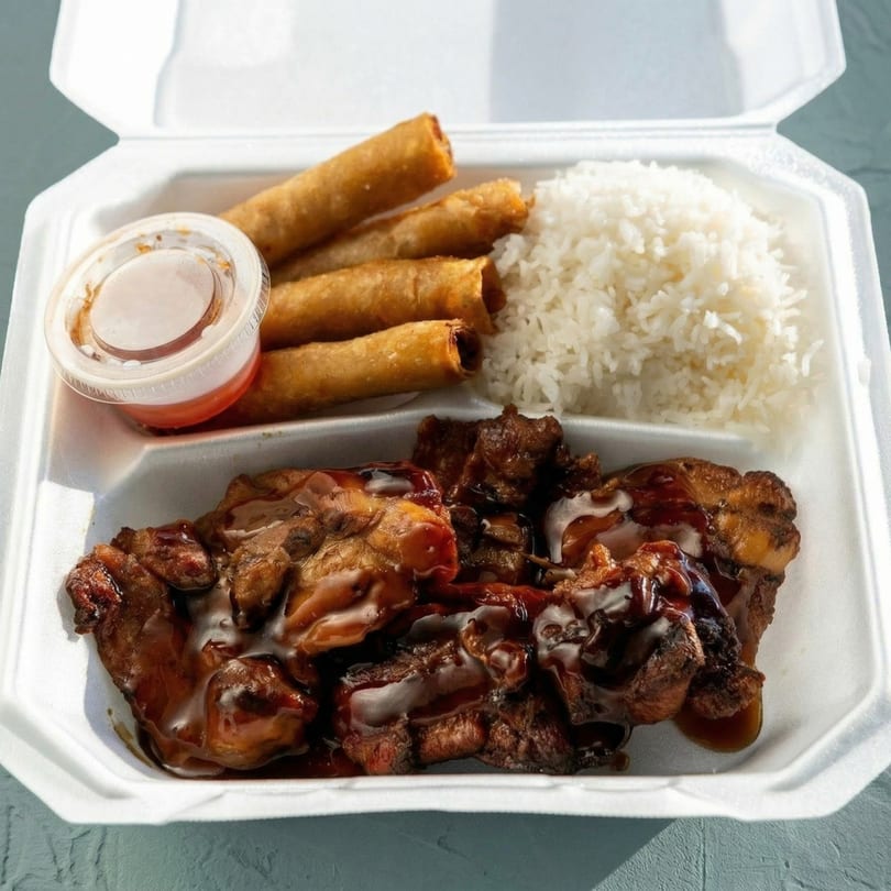 A takeout container with saucy chicken, white rice, four egg rolls, and a small cup of sauce.