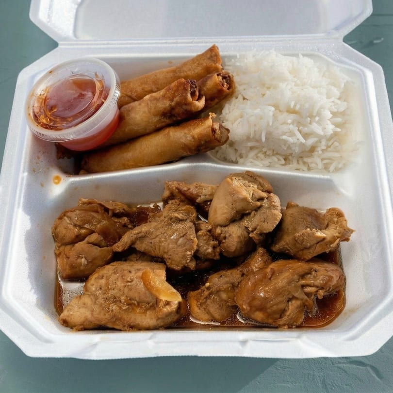 A takeout container with chicken adobo, white rice, three lumpia rolls, and a side of sweet chili sauce.